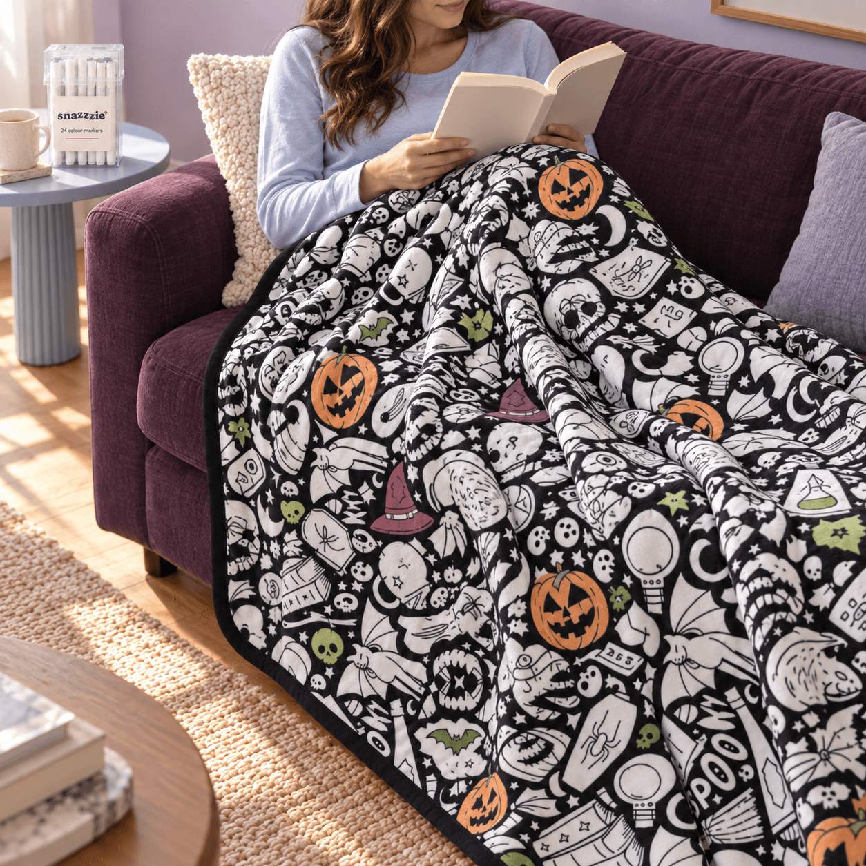 Una donna legge su un divano color prugna sotto una coperta Brividi di Halloween parzialmente colorata, con l’astuccio dei pennarelli sul tavolino